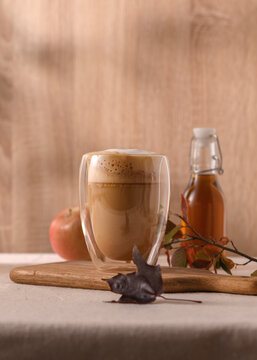 Foamy apple maple latte in a clear glass on a wooden board, apple and syrup bottle in the background, autumn leaves creating cozy mood.