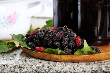 Ripe mulberries are stacked on a wooden tray, with two glass bottles filled with dark liquid next...