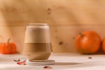 Creamy pumpkin latte in a clear glass stands on a light table with autumn leaves, cozy pumpkins, and soft sunlight in the background.