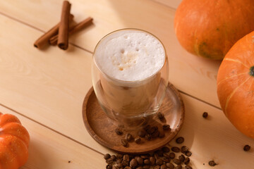 Autumn latte with airy foam in a double glass on a wooden plate, surrounded by coffee beans, pumpkins, and cinnamon sticks in warm light.