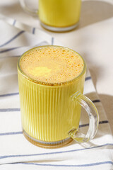 Close-up of golden turmeric latte with frothy texture in glass cup on striped linen, warm drink for cozy mornings and relaxation.