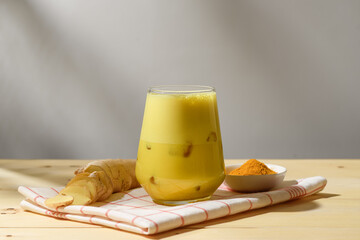 Cold golden turmeric latte in transparent glass with ice cubes, fresh sliced ginger root and turmeric powder on checked cloth, natural wellness drink.