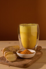 Glass with frothy golden turmeric latte on wooden board with sliced ginger root and turmeric powder, warm brown background creates cozy health vibe.