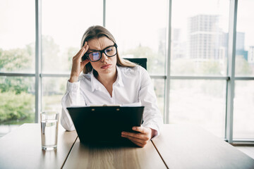 Focused businesswoman reviewing documents at a modern office workspace with glasses, fostering...