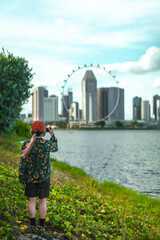 Front view of a woman standing with her back to a green grassy waterfront bank, taking pictures at the Singapore Flyer in Singapore. In the background, there are trees and buildings in the evening.