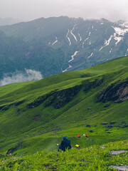 Photo from the back of a man hiker in a raincover sitting at a high mountain pass surrounded by dramatic clouds and rocky alpine terrain. Idea for a solo trekking adventure in the Himalayas.