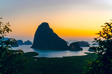 Numerous limestone mountains in the bay in Phang Nga Province, Thailand. The light of the sky is colorful of rising sun in the early morning. There is a blurred foreground of grass on the left.