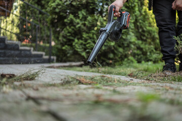 Person cleaning garden path with a cordless electric blower. Dust and grass clippings scatter across the pavement. Concept of outdoor maintenance and garden tools