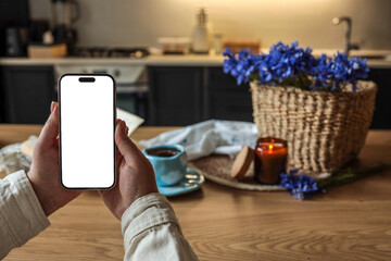 Hands holding smartphone with blank white screen in cozy kitchen setting. Cup of coffee, candle,...