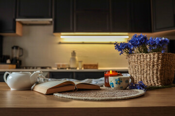 Cozy kitchen scene with a teapot, open book, lit candle, and floral cup on a wooden table. A woven basket of cornflowers adds a rustic charm to the warm, homey atmosphere