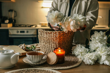 Person arranging fresh white peonies into a woven basket on a wooden kitchen table. Nearby are a...