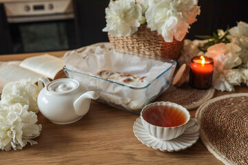 Cozy breakfast scene with a cup of tea, teapot, cinnamon rolls in a glass dish, open book, burning candle, and white peonies on a wooden table in a warm kitchen setting