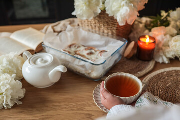 Warm cozy scene with a person holding a tea cup, cinnamon rolls in a glass tray, a teapot, open book, candle, and blooming white peonies on a wooden table in soft light