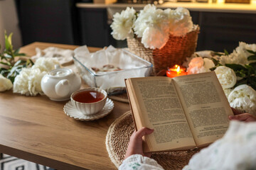 Open book on a wooden table beside a cup of tea, candle, and peonies in a cozy kitchen setting....