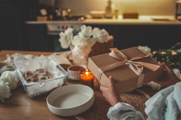 Hands holding a kraft gift box with satin ribbon in cozy kitchen setting. Cinnamon rolls,...