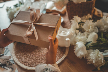 Hands holding a kraft gift box with satin ribbon in cozy kitchen setting. Cinnamon rolls, candlelight, and white peonies create a warm and inviting atmosphere on wooden table