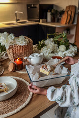 Child serving fresh cinnamon roll from glass baking dish in cozy kitchen. Rustic breakfast scene with flowers, candlelight, teapot, and warm natural textures