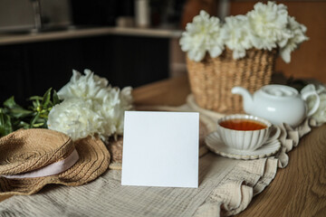 Blank white square card mockup on cozy rustic table with straw hat, cup of tea, teapot, wicker basket of white peonies, and linen fabric. Styled warm countryside scene