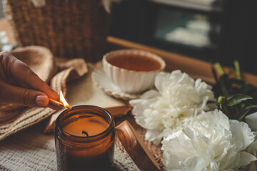 Hand lighting a candle in a cozy flat lay scene with tea, white peonies, straw hat, and an open book. Warm summer moment of relaxation and self-care in soft natural light
