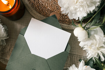 Minimalist flat lay with blank white card in green envelope, white peonies, candlelight and natural textures. Warm rustic background for romantic message, wedding or branding mockup