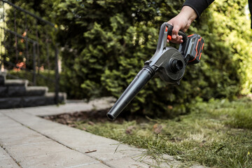 Person cleaning garden path with a cordless electric blower. Dust and grass clippings scatter across the pavement. Concept of outdoor maintenance and garden tools © shine.graphics