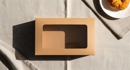 A tan box with a window sits on a linen tablecloth next to a plate with a pastry on it in sunlight