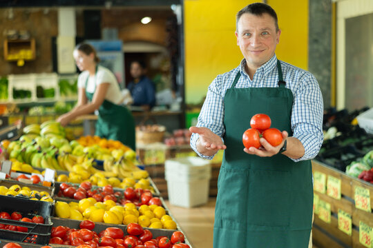 With practiced hand, man supermarket worker arrange tomatoes in neat pyramid by window. Male seller of vegetable department of store replenishing showcase with tomato, puts ones on showcase