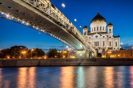 View of the Cathedral of Christ the Saviour glowing warmly under a twilight sky, framed by the modern steel bridge and reflected in the river, Moscow, Moscow, Russia.