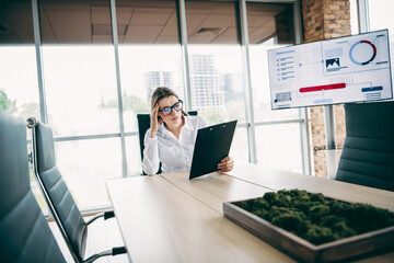 Busy businesswoman reviewing documents in a modern office setting with analytical charts visible on...