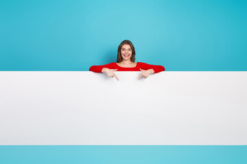 Young cheerful woman pointing down against a blue background showcasing a blank white panel for...