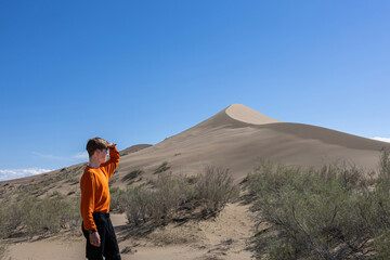 A young man in an orange sweater looking at a large sand dune in the desert under a clear blue sky