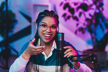 Young African American woman recording a podcast at home studio with microphone under colorful neon...