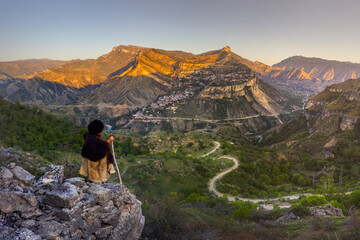 Dagestan, Russia - 23 July 2025: View of a solitary figure overlooking the mountainous landscape, where golden light kisses the rugged peaks and a winding road snakes through the verdant valley below.