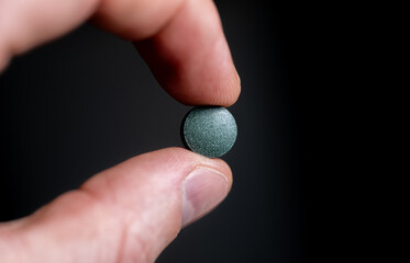 Single green round tablet held between fingers on dark background. Dietary supplement and...