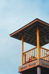 A wooden beach guard tower stands tall against a clear blue sky, featuring a red object inside.