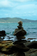 A stack of balanced rocks stands on a shore, with the ocean and distant mountains visible in the background.