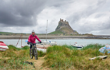 nice senior woman riding her electric mountain bike on Holy Island, with medieval Lindisfarne castle in background, Northumbria, England, UK