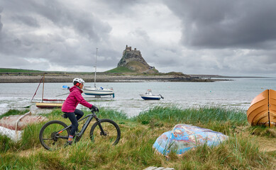 nice senior woman riding her electric mountain bike on Holy Island, with medieval Lindisfarne castle in background, Northumbria, England, UK