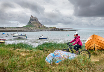 nice senior woman riding her electric mountain bike on Holy Island, with medieval Lindisfarne castle in background, Northumbria, England, UK