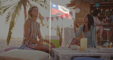 Laughing two women holding cocktails on pallet table at beach sunset, with tiki bar behind