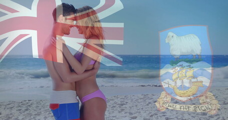 Embracing couple in swimwear standing at water's edge on beach, Union Jack and Falklands overlays
