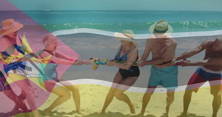 Pulling five retired friends wearing swimwear leaning back on sandy beach, with tug-of-war rope