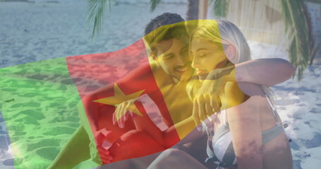 Sitting couple in swimwear embracing each other on sandy beach near calm sea, with flag overlay