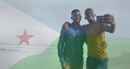 Smiling friends taking selfie on sandy shoreline with Djibouti flag overlay, using smartphone