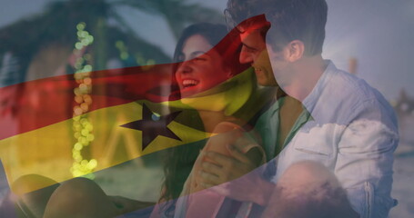 Smiling couple sitting on sandy shore at dusk, with translucent flag overlay and string lights