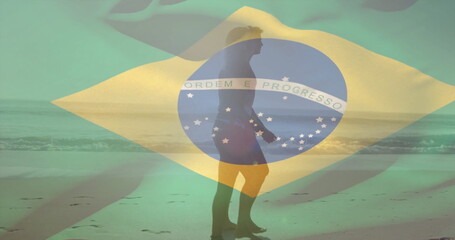 Walking surfer in board shorts carrying surfboard along beach at low tide, Brazilian flag overlay