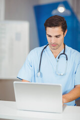Professional wearing scrubs and stethoscope typing on silver laptop on white desk in nurse station
