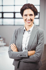 Businesswoman wearing grey suit posing crossing arms at desk with monitor and grid window in office