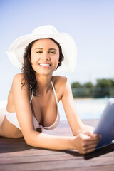 White sunhat lying next to tablet on wooden pool deck with sunlit water and greenery