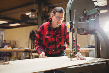 Mature man wearing flannel shirt guiding plywood through vertical band saw in woodshop with sawdust